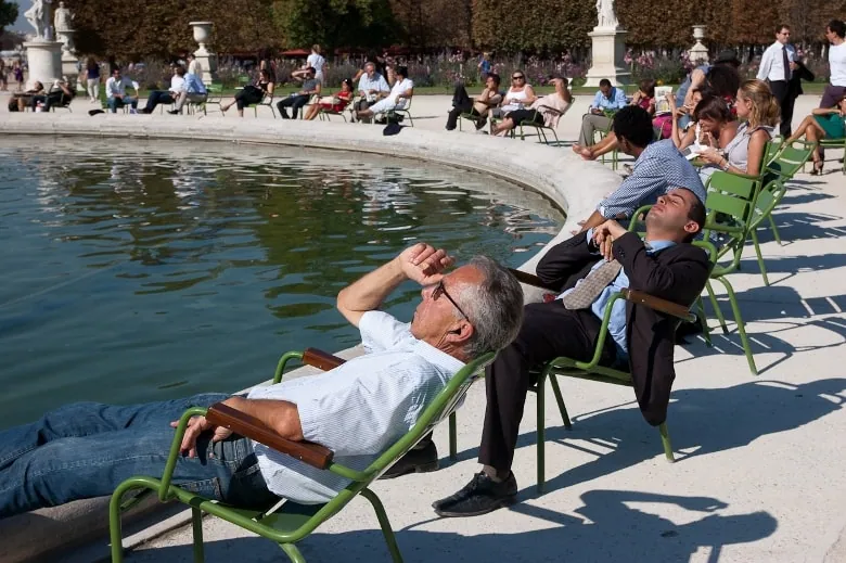 Homem em fato a descansar numa cadeira de jardim ao sol junto a um lago.